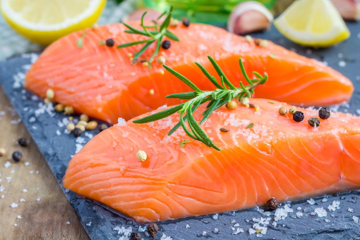 Two pieces of raw salmon topped with salt, pepper, and rosemary. Sitting on a grey board with lemon and garlic.